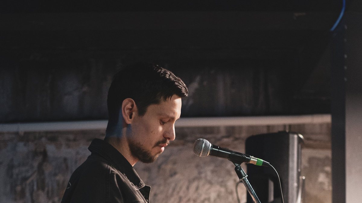 Man in a denim jacket looking down as he reads aloud into a stage microphone