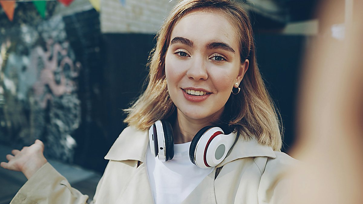 Smiling young woman with white headphones around her neck talking with someone outdoors