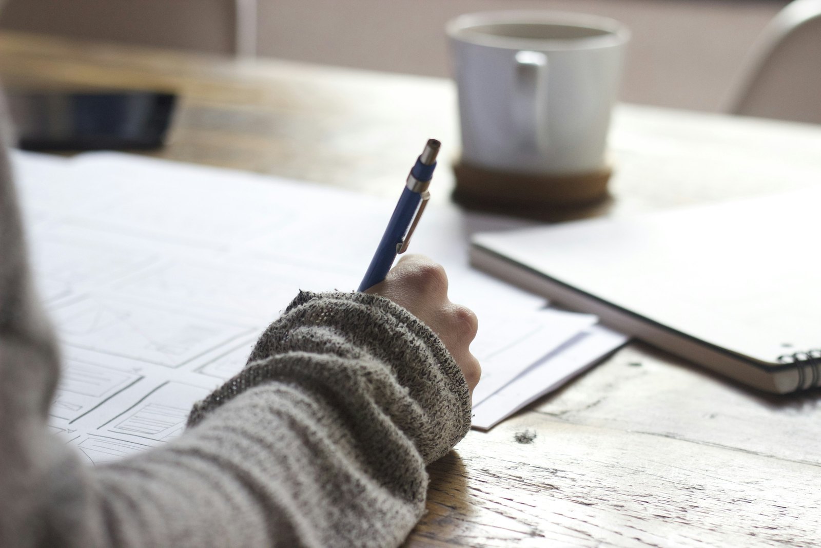 Person writing study notes at a wooden table beside a mug